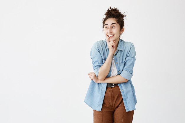 Femme aux cheveux noirs émotionnelle positive avec chignon souriant largement, gardant le doigt sur les dents, regardant de côté, pensant à quelque chose de bien. Fille excitée en chemise en jean pose contre le mur blanc