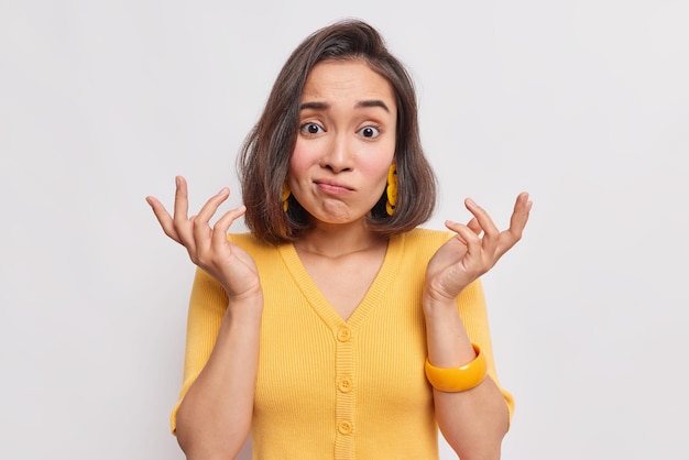 femme aux cheveux noirs expression désemparée lève les mains hausse les épaules avec étonnement ne sait pas comment agir porte un pull jaune décontracté isolé sur blanc