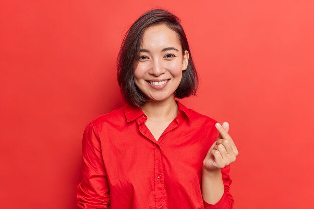 femme aux cheveux noirs fait un mini geste de coeur symbole coréen de l'amour exprime sa sympathie vêtue d'une chemise rouge dans un ton avec