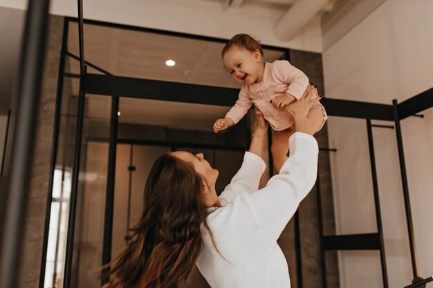 Femme aux cheveux noirs en peignoir blanc joue avec sa fille et la jette dans l'appartement.