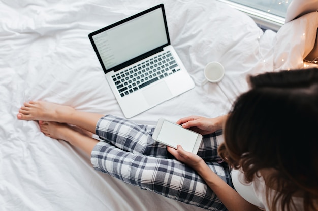 Femme aux cheveux noirs raffinée à l'aide de téléphone assis sur le lit. Freelance féminine travaillant avec un ordinateur portable à la maison le matin.