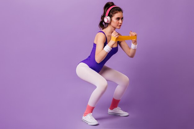 Femme aux cheveux noirs en survêtement violet et baskets blanches accroupie avec bande pour le sport