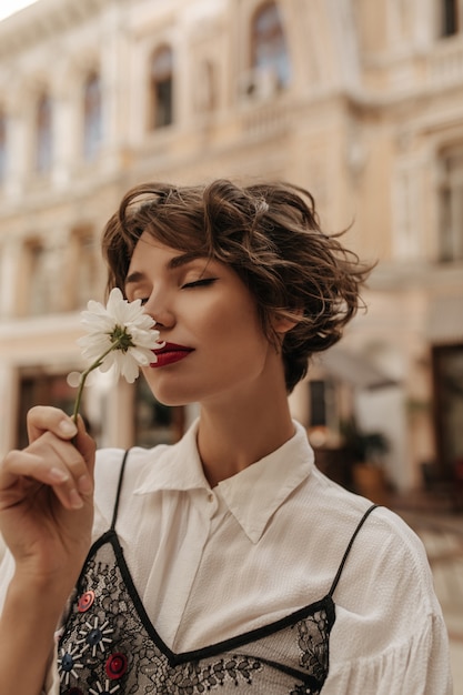 Photo gratuite femme aux cheveux ondulés en chemise légère avec fleur reniflante de dentelle noire en ville. femme tendre aux lèvres rouges et cheveux courts pose à la rue.
