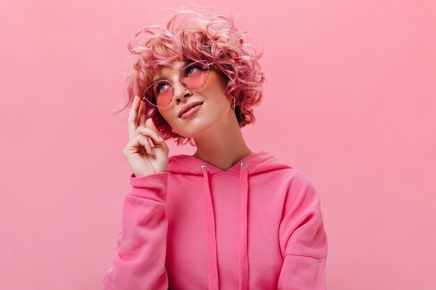 Femme aux cheveux roses bouclés et rêveurs dans un sweat à capuche surdimensionné lumineux sourit sur une charmante fille isolée dans des lunettes de soleil pose sur fond rose