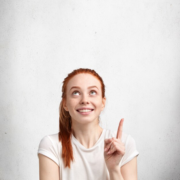 Femme aux cheveux rouge positif avec un sourire agréable, habillé avec désinvolture, pointe vers le haut au plafond, annonce quelque chose à l'espace copie vierge