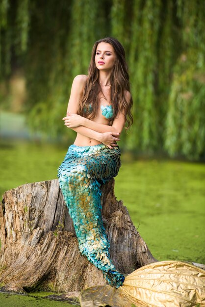 femme aux longs cheveux bruns et habillée comme une sirène assise sur la pierre au-dessus de l'eau