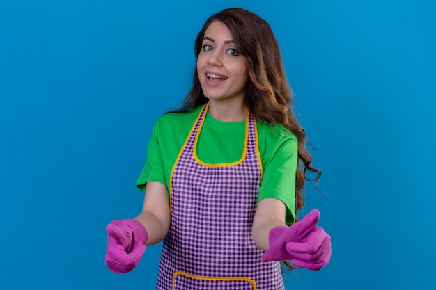 Femme aux longs cheveux ondulés portant un tablier et des gants en caoutchouc souriant avec visage heureux montrant les pouces debout sur le bleu