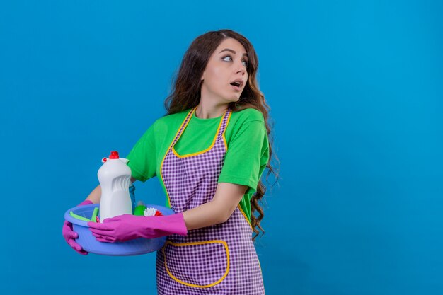 Femme aux longs cheveux ondulés portant un tablier et des gants en caoutchouc tenant un bassin plein d'outils de nettoyage à côté avec surprise face peur et sorti debout sur bleu