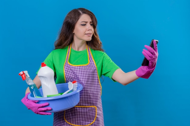 Photo gratuite femme aux longs cheveux ondulés portant un tablier et des gants en caoutchouc tenant un bassin plein d'outils de nettoyage à la recherche de téléphone mobile avec une expression confuse debout sur le bleu