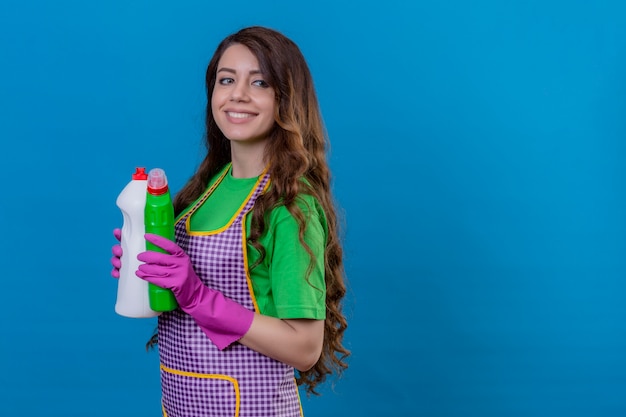 Femme aux longs cheveux ondulés portant un tablier et des gants en caoutchouc tenant des bouteilles avec des produits de nettoyage souriant debout confiant sur bleu