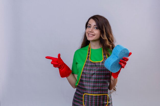 Femme aux longs cheveux ondulés portant un tablier et des gants en caoutchouc tenant une éponge et pointant avec l'index sur le côté souriant debout amical