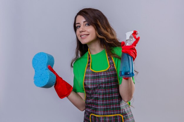 Femme aux longs cheveux ondulés portant un tablier et des gants en caoutchouc tenant une éponge et un spray de nettoyage souriant debout amical