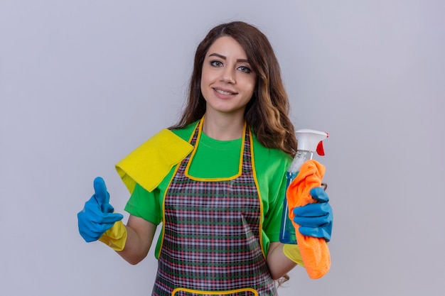 Femme aux longs cheveux ondulés portant un tablier et des gants en caoutchouc tenant un tapis et un spray de nettoyage à la recherche d'un sourire confiant et positif montrant les pouces vers le haut prêt à nettoyer debout sur bleu