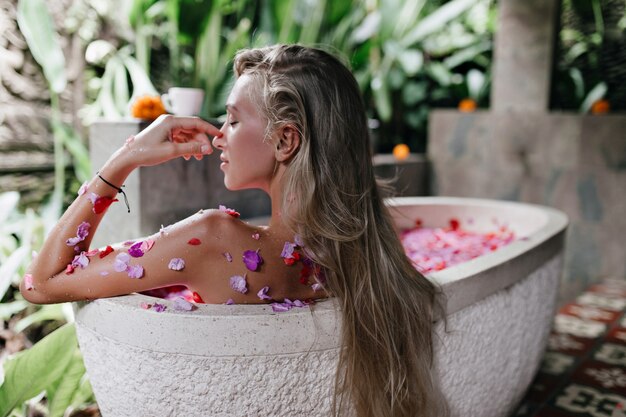 femme aux longs cheveux raides assis dans une baignoire pleine de pétales de rose. Plan intérieur d'une magnifique femme bronzée se reposant à la maison et faisant du spa.