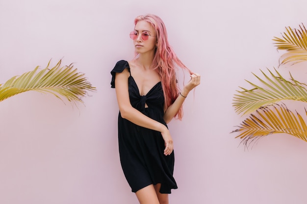 femme aux longs cheveux roses debout à côté de palmiers exotiques en studio.