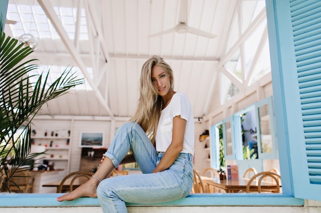 Femme aux pieds nus inspirée avec un joli sourire posant dans un café. Photo du modèle féminin blond mignon avec l'expression du visage excité assis sur le rebord de la fenêtre.