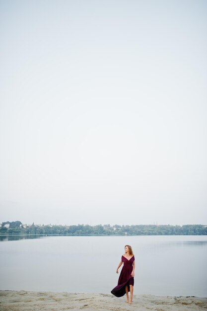 Femme aux pieds nus sensuelle blonde en robe marsala rouge posant contre le lac sur le sable