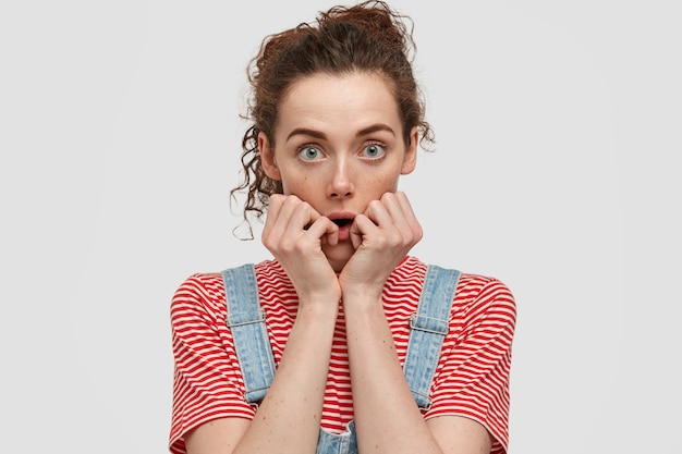 La femme aux taches de rousseur aux yeux verts garde les mains près de la bouche, a l'air perplexe, a les cheveux bouclés foncés, porte un t-shirt à rayures décontracté avec une salopette en denim, isolée sur un mur blanc. Les gens et l'émerveillement