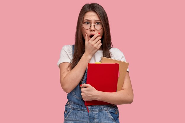 Une femme aux taches de rousseur émotive couvre la bouche, a une scène effrayante, porte un bloc-notes, porte un t-shirt blanc décontracté et une salopette