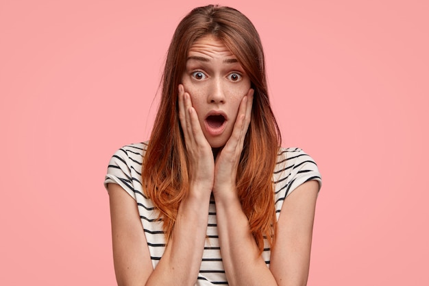 Femme aux taches de rousseur avec garde la mâchoire tombée alors qu'elle réalise quelque chose de surprenant, porte un t-shirt à rayures décontracté contre un mur rose