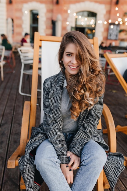 Femme aux yeux bleus bien habillée avec une coiffure frisée posant avec le sourire sur un fauteuil inclinable. Fille blanche élégante en jean bleu assis dans une chaise longue au café, en attendant des amis.