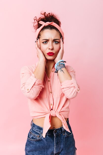 Femme aux yeux bleus en chemise pin-up et jeans regarde la caméra avec inquiétude. Fille bouclée avec des lèvres rouges posant sur un espace rose.