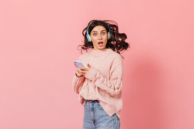 Une femme aux yeux bleus regarde avec surprise la caméra sur fond rose. Une fille en pull tricoté écoute de la musique dans des écouteurs et tient un smartphone