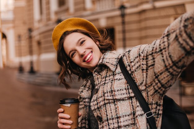 Femme aux yeux bruns en béret orange fait selfie contre le mur de la belle maison