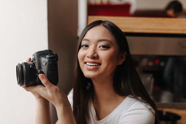 Femme aux yeux bruns en haut blanc sourit et tient devant tout en posant dans son appartement