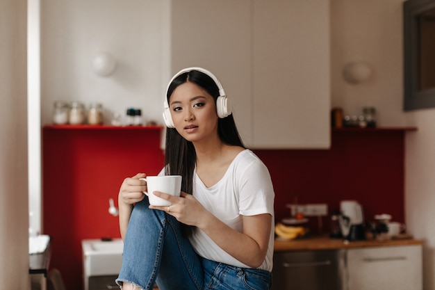 Femme aux yeux bruns en T-shirt blanc et écouteurs massifs regarde à l'avant, posant avec tasse sur fond de cuisine