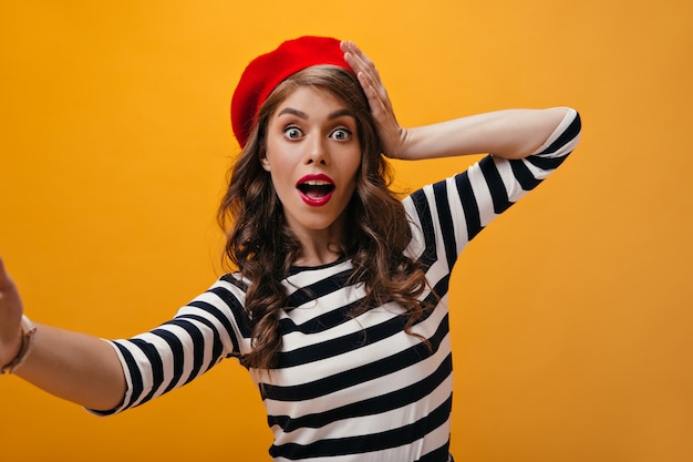 Femme aux yeux ouverts en béret rouge fait selfie. Surpris fille bouclée avec chapeau lumineux en chandails modernes à rayures regardant dans la caméra.