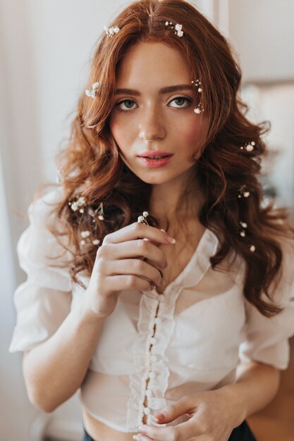 Femme aux yeux verts avec des fleurs dans les cheveux ondulés se penche sur la caméra. Instantané de femme en chemisier blanc.