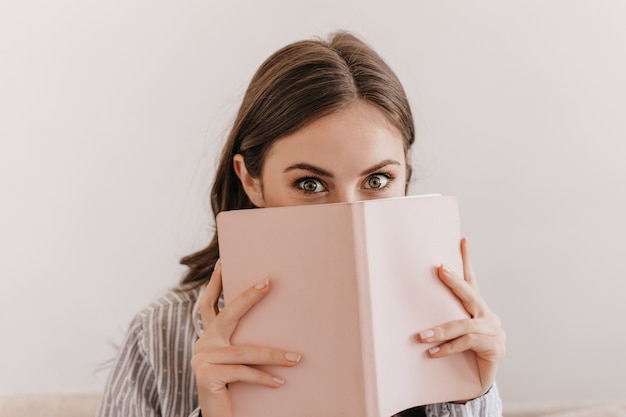 Femme aux yeux verts en pyjama rayé regarde à l'avant et se couvre le visage avec un ordinateur portable