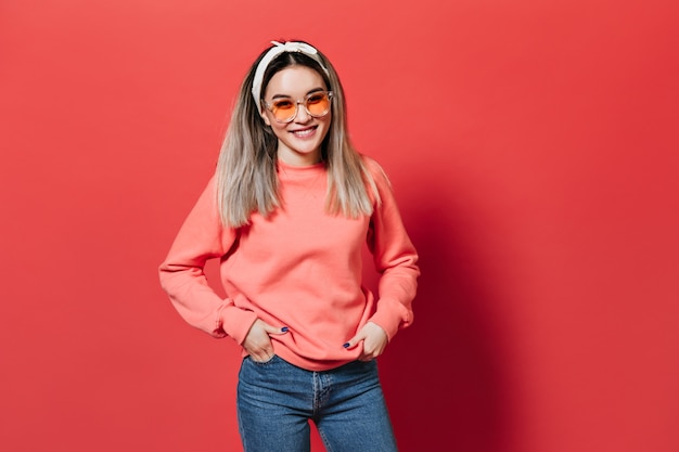 femme avec bandeau, portant des lunettes orange et un sweat-shirt, posant sur un mur rouge