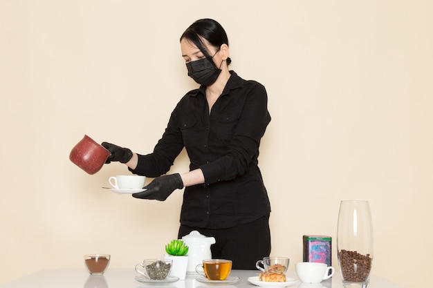 Femme barista en pantalon chemise noire avec du café brun thé séché ingrédients de l'équipement en masque stérile noir sur le mur blanc