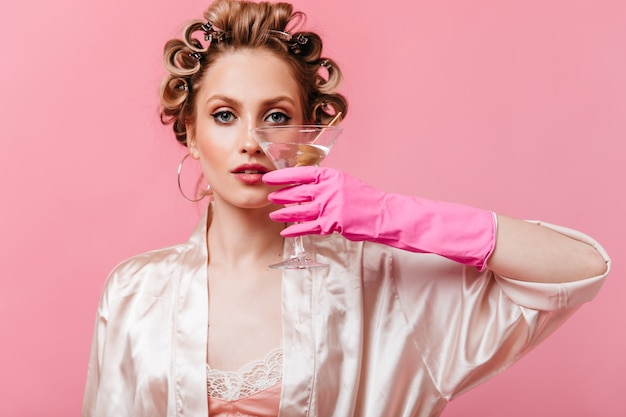 Femme en bigoudis pose avec verre à martini sur mur rose