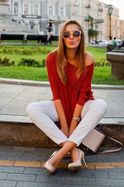 Femme blanche à la mode en pull assis dans la rue dans le centre-ville. Lunettes de soleil élégantes. Ambiance d'automne.