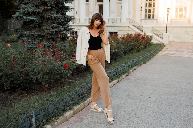 Femme blanche à la mode en veste posant dans la rue. Poils ondulés, maquillage naturel. Longueur totale.