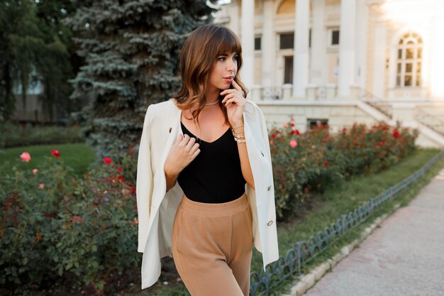 Femme blanche à la mode en veste posant dans la rue. Poils ondulés, maquillage naturel.