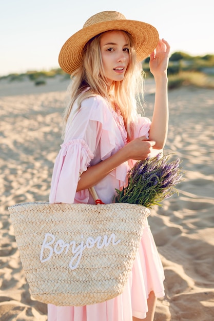 Femme blanche romantique au chapeau à la mode et élégante robe rose posant sur la plage. Sac de paille et bouquet de fleurs.