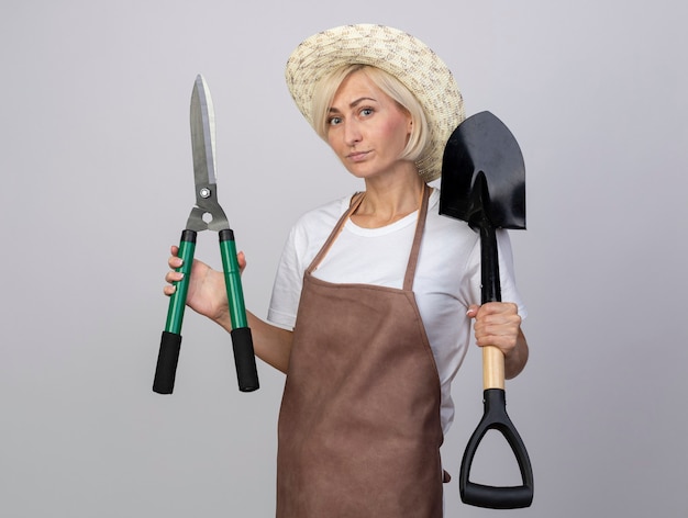 Photo gratuite femme blonde d'âge moyen confiante en uniforme de jardinier portant un chapeau tenant des ciseaux à haie et une pelle