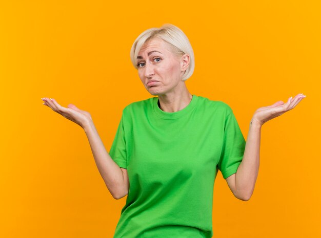 Femme blonde d'âge moyen confus à l'avant faisant je ne sais pas de geste isolé sur mur jaune