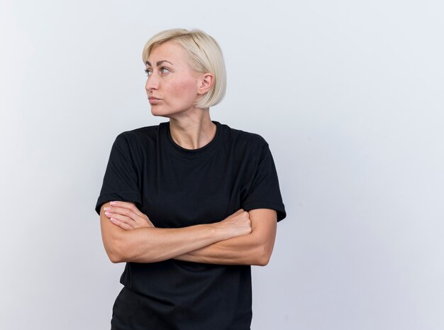 Femme blonde d'âge moyen réfléchie debout avec une posture fermée à côté isolé sur mur blanc
