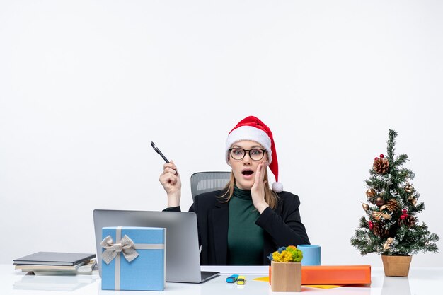 Femme blonde agitée avec un chapeau de père Noël assis à une table avec un arbre de Noël et un cadeau sur elle sur fond blanc