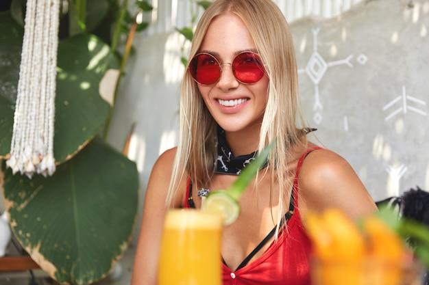 Femme blonde assez heureuse dans des lunettes de soleil rouges élégantes, recrée dans un restaurant confortable avec un cocktail orange frais
