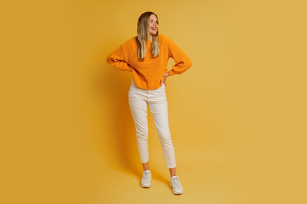 Femme blonde au visage suprice en pull d'automne élégant orange posant sur jaune. Longueur totale.