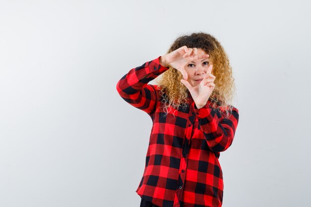 Femme blonde aux cheveux bouclés faisant un geste de cadre en chemise à carreaux et regardant concentré, vue de face.