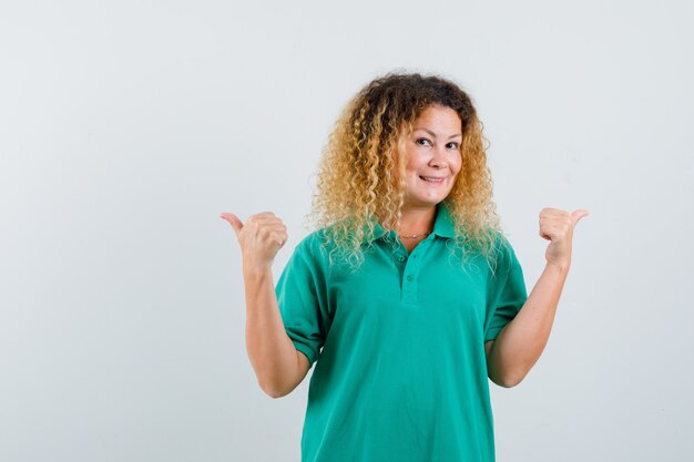 Femme blonde aux cheveux bouclés pointant vers les directions opposées en T-shirt vert et semblant joyeuse. vue de face.