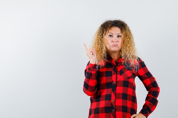 Photo gratuite femme blonde aux cheveux bouclés pointant vers le haut en chemise à carreaux et l'air déçu, vue de face.