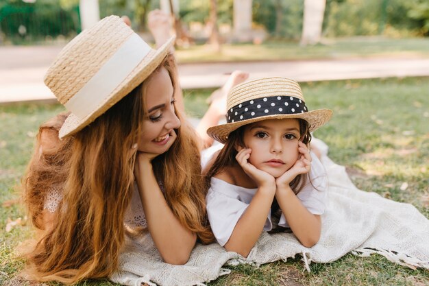 Femme blonde aux cheveux longs en canotier à la mode avec le sourire à la petite fille pensive. Charmante fille au chapeau de paille avec un ruban noir étayant le visage avec les mains tout en se détendant sur l'herbe après le match.